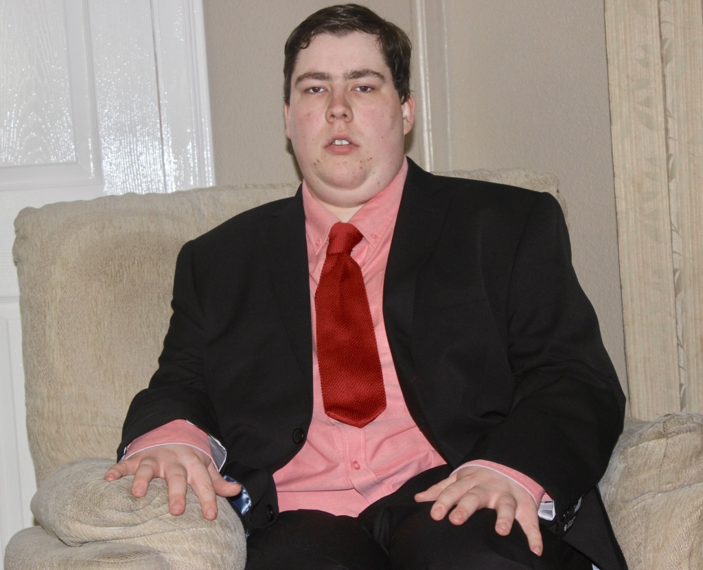 Me sat on a beige chair, wearing a black suit jacket, a pink shirt, and a red tie, on a grey background.