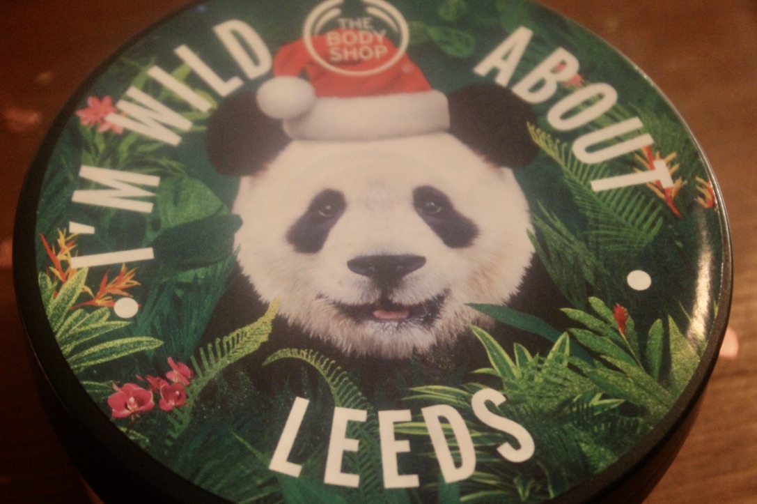 A Green body butter tub, with a label with white font and a picture of a Panda on it, on a wooden background.