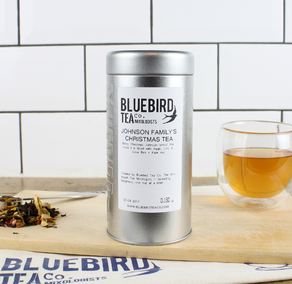 A metallic cylindrical tin, with a white label, next to a glass mug containing golden tea, on a white background.
