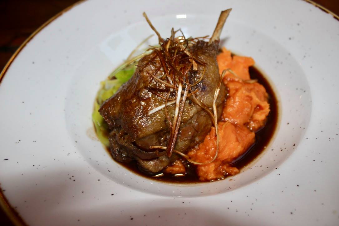 Some dark Duck meat, dark green leeks, bright orange Sweet Potato Mash, and some dark brown gravy in a white bowl, on a grey background.
