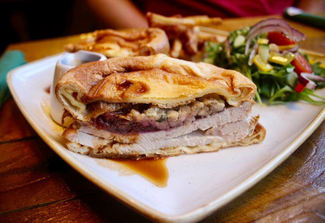 A golden Yorkshire pudding with a pale Turkey filling, and a slightly golden Yorkshire Pudding underneath it, some light brown gravy, dark golden chips, and a colourful salad, on a dark background.