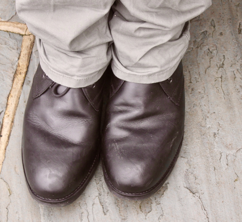 A person wearing a pair of Black leather boots pressed together, with a hint of green stretched straight chinos, on a stone background.