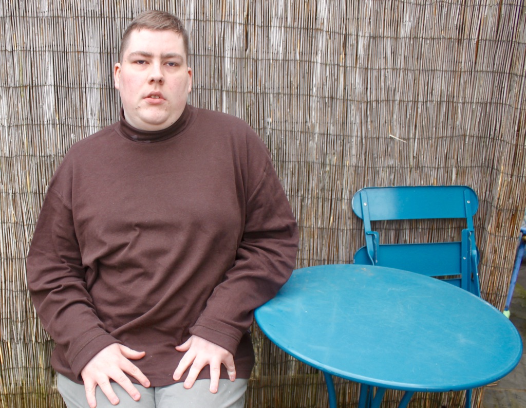 A person wearing a chocolate brown roll neck top, sat down on a blue chair next to a blue table, on a light wood background.
