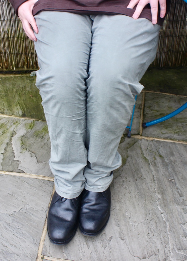 A person wearing a pair of Green stretched straight chinos with a pair of black leather boots, sat down on a blue chair, against a stone floor with a light wood background.