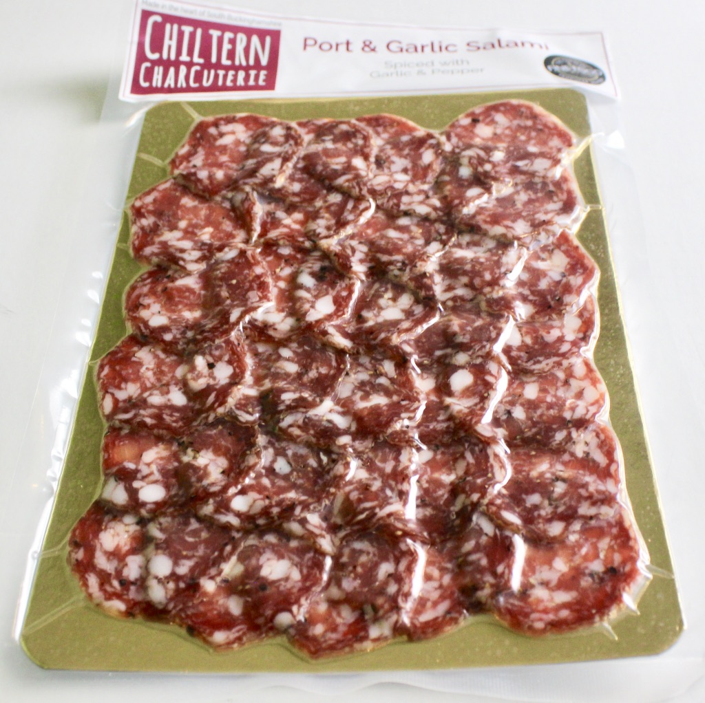 Rectangular see through packet with a red, white and blue Union Jack flag stuck on top containing Dark red and white Chorizo slices with a silver label containing a red logo saying Chiltern Charcuterie and Port & Garlic Salami with a description in black writing on a white background