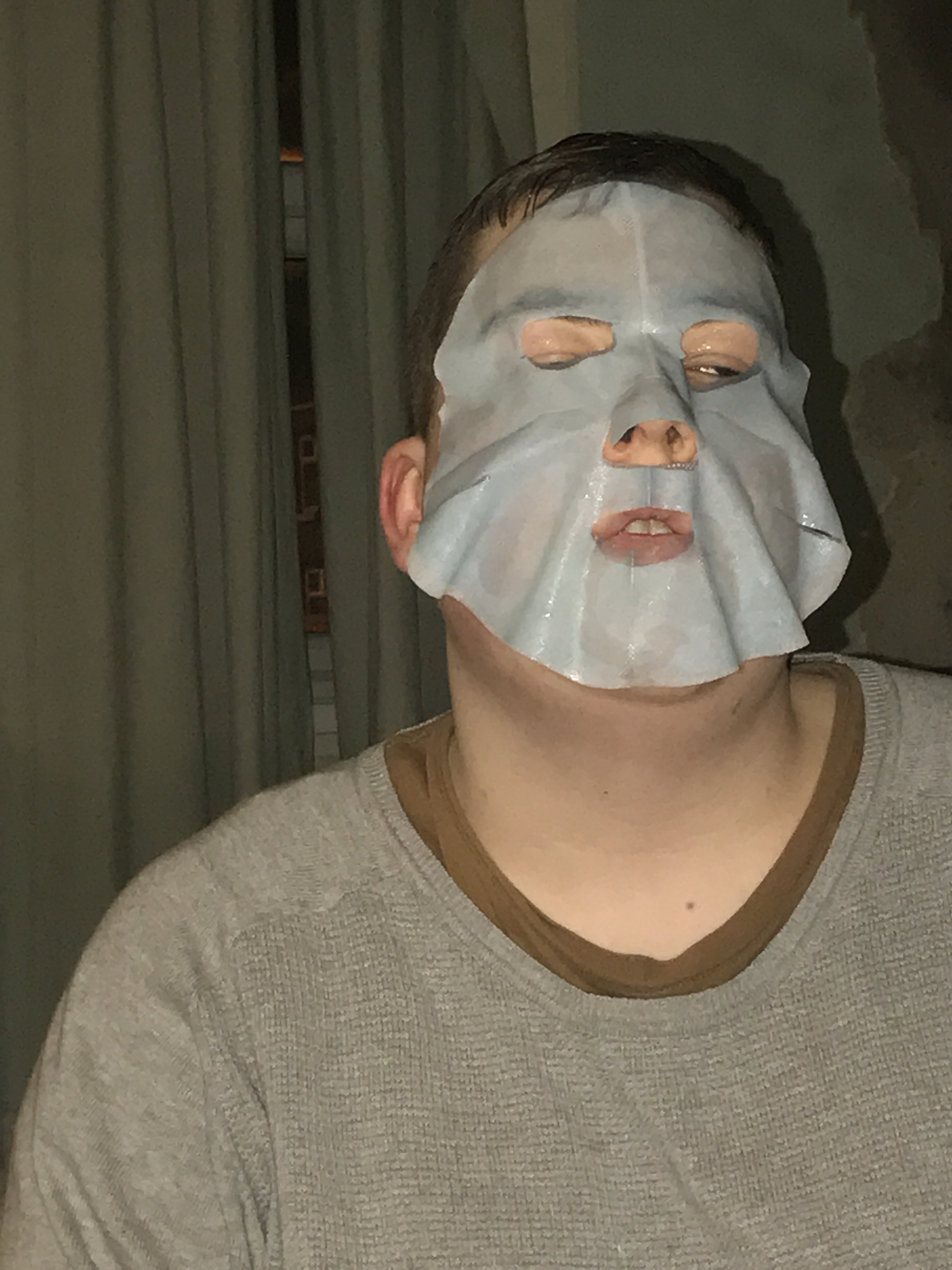 A person wearing a grey jumper and a light blue sheet mask on a dark background.