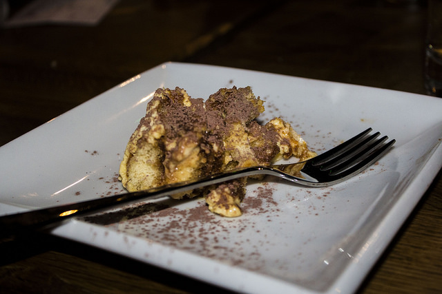 A layered Tiramisu with layers of brown, white and black, on a white square plate with a dark silver fork, on a dark background.