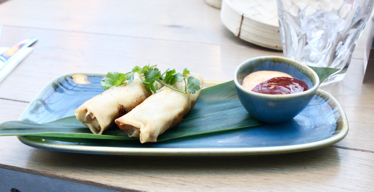 Some long tan coloured Spring Rolls resting on a large green leaf next to a dark oval pot full of red ketchup and white mayonnaise on a black rectangular plate on a tan table, on a white background.