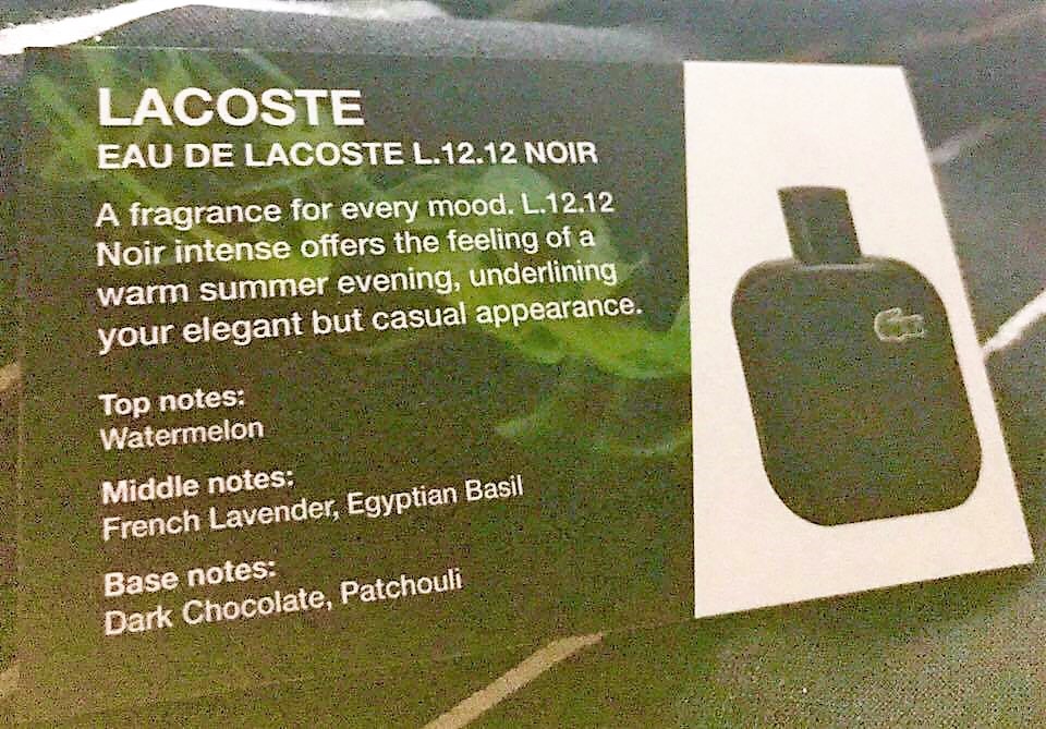 A small Gold rectangular piece of card with a description of the Lacoste Eau De Lacoste L.12.12 Noire Eau De Toilette fragrance written in white writing next to a black square bottle, on a black background