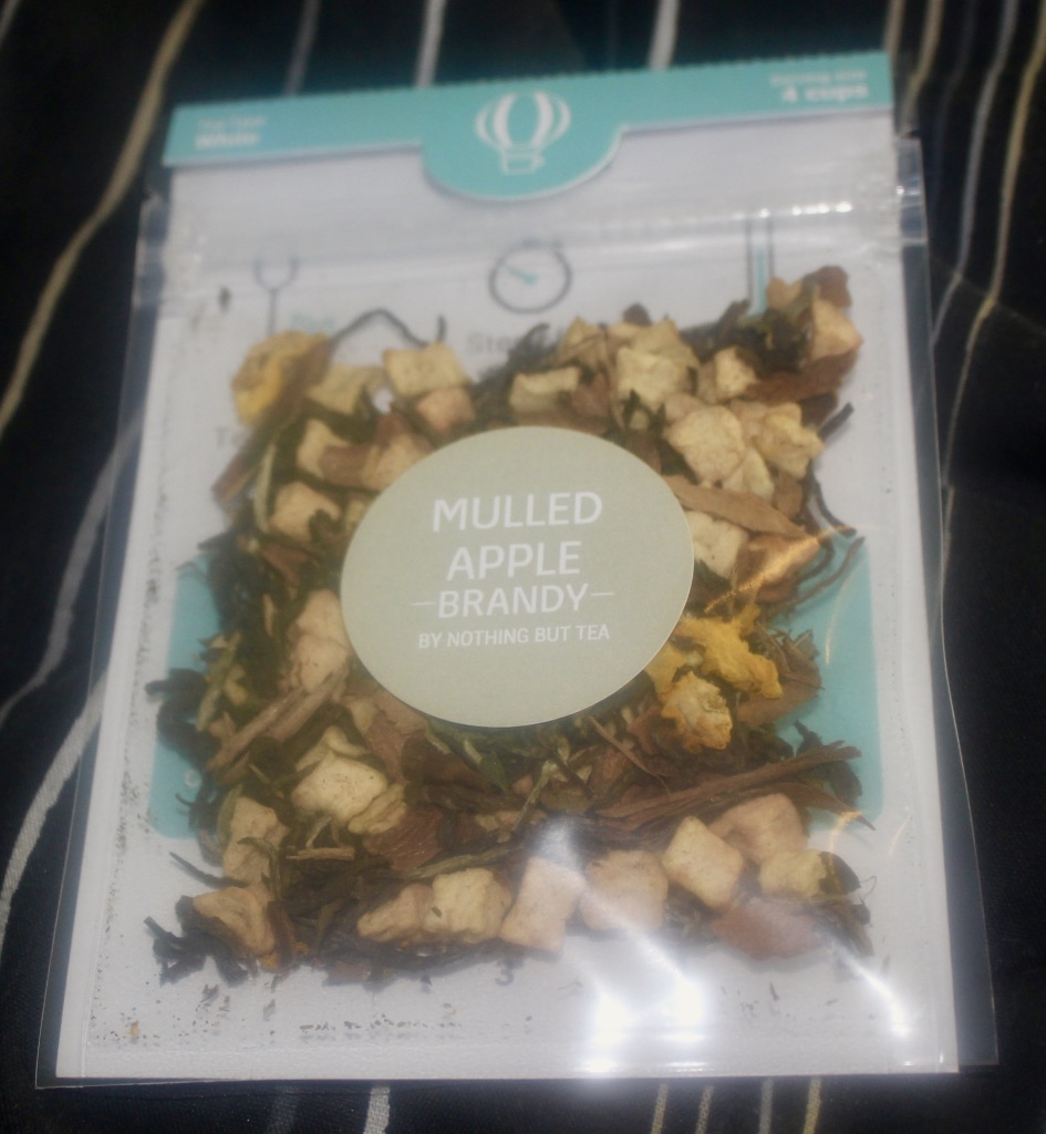 A silver pouch with a large silver circular sticker that has Nothing But Tea Mulled Apple Brand Tea written on it, that contains brown tea leaves and some pieces of green Apple, on a black background.