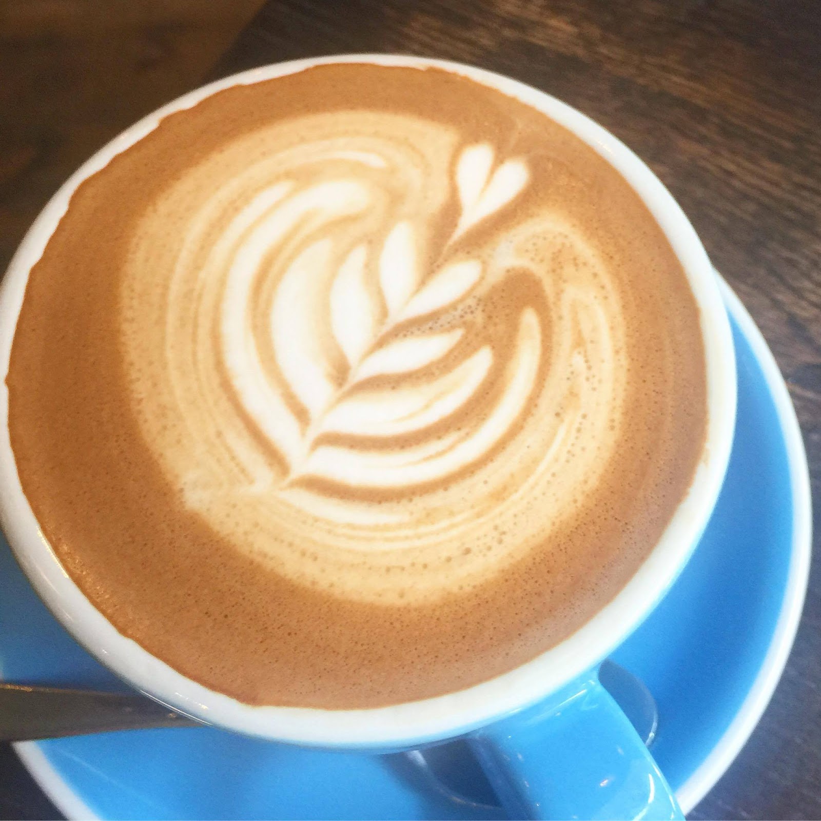 A light brown coffee with white ribbon detailing on the top of it, in a blue round cup and saucer, on a dark background.