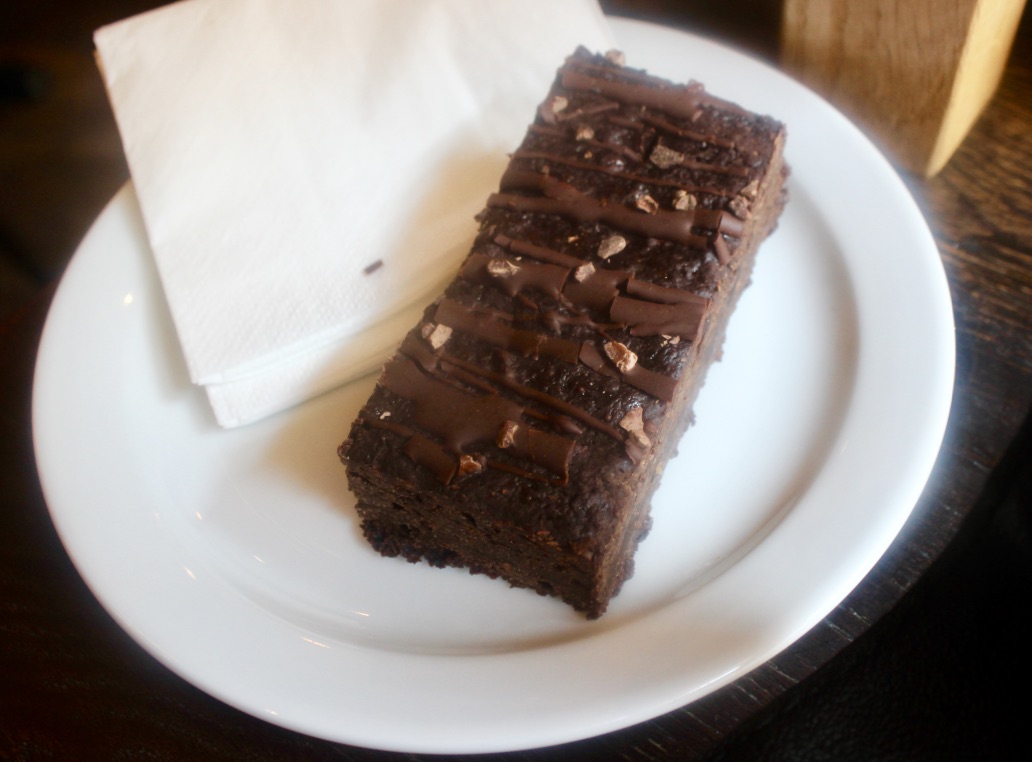 A long thick rectangular shaped dark brown chocolate brownie with some flex of dark brown nuts, on top of a small rectangular white tissue, and a small circular white plate, on a dark background.