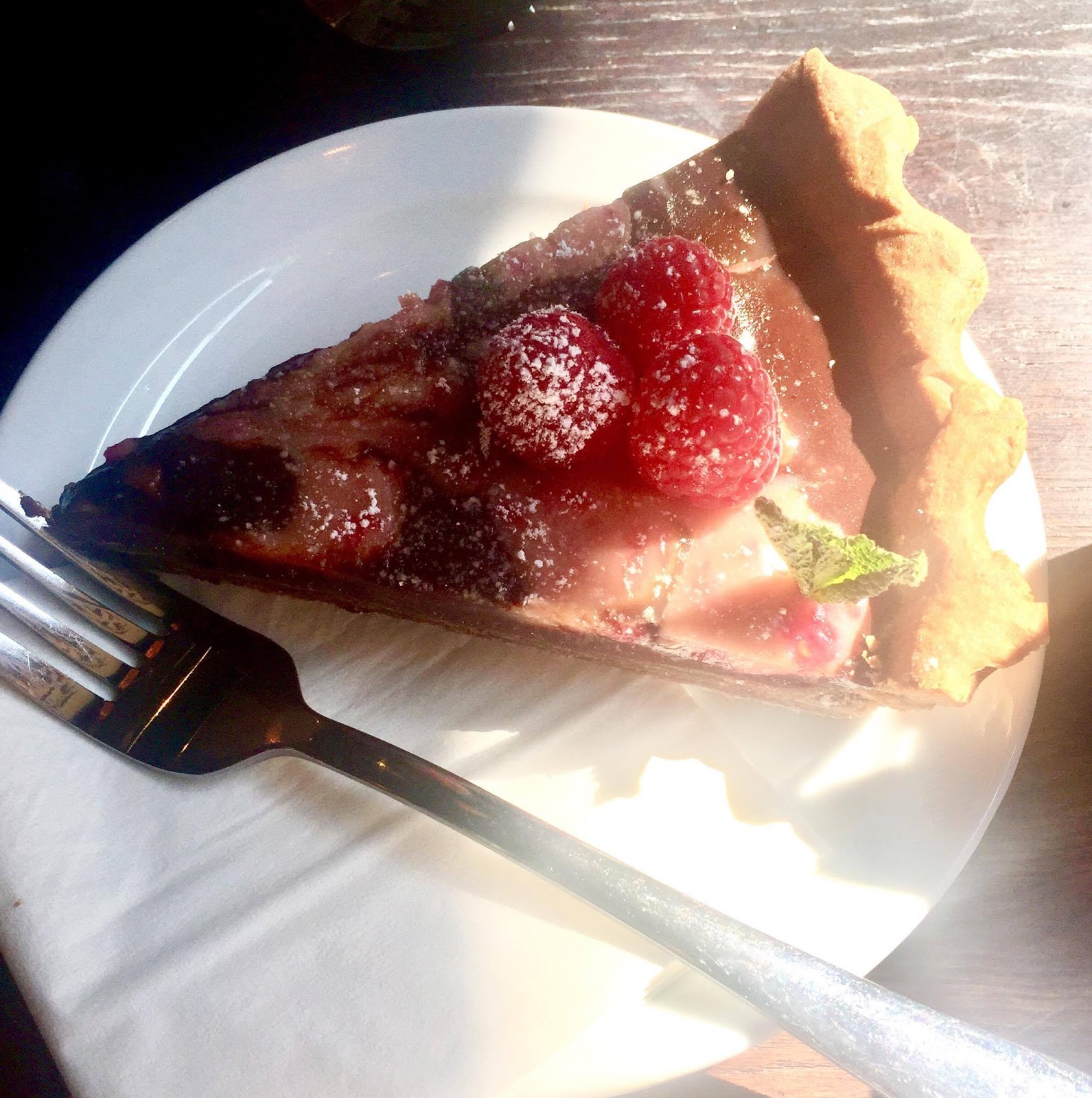 A large wedge of tart consisting of some light golden brown pastry, some red Raspberry coulis, and some dark brown chocolate ganache, all topped off with red raspberries, on a circular small plate, on a light brown background.