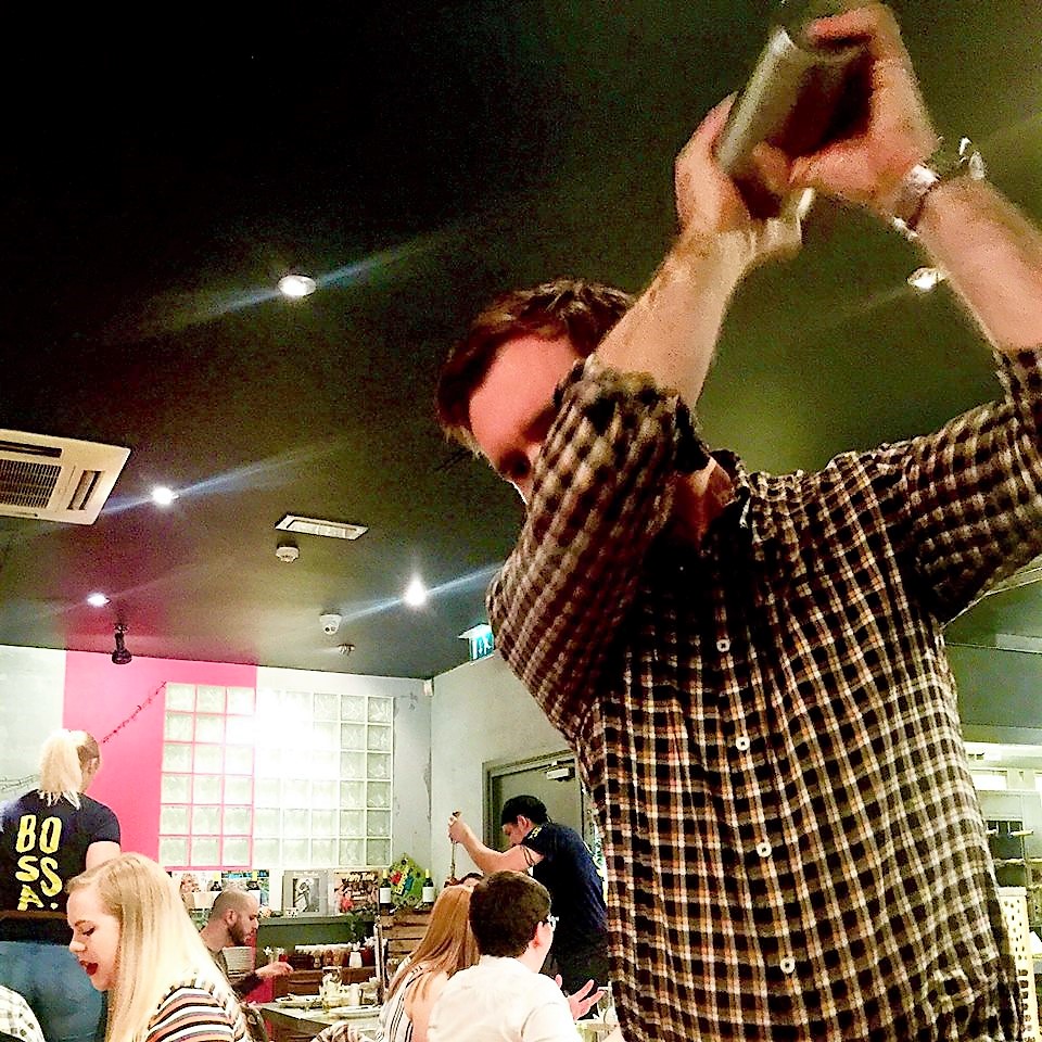 A tall white man in a white and grey chequered shirt holding a metal cocktail shaker, on a dark background.