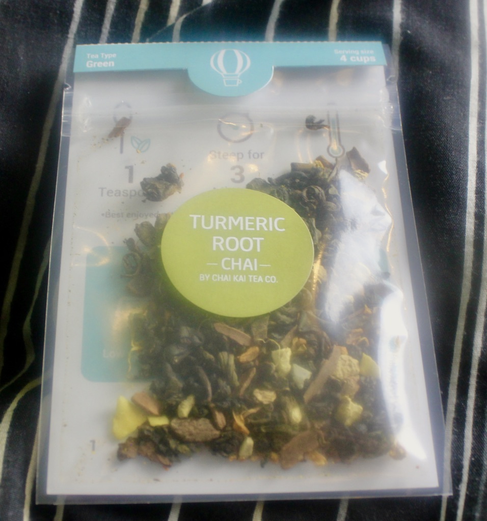 A silver pouch with a large green circular sticker that has Chai Kai Tea Company Turmeric Chai Tea written on it, that contains some brown leaves and some brown spices, on a dark background.