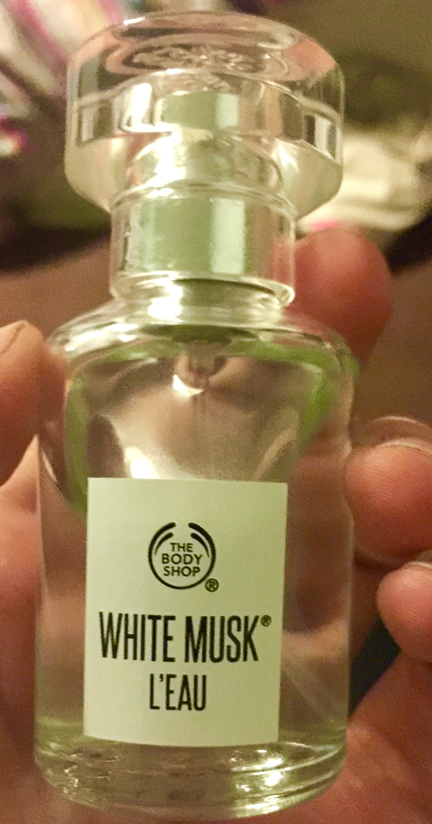 A tall glass bottle filled with some clear liquid with a bright white rectangular label that has the body shop white musk written on it in bold black writing, on a light background.