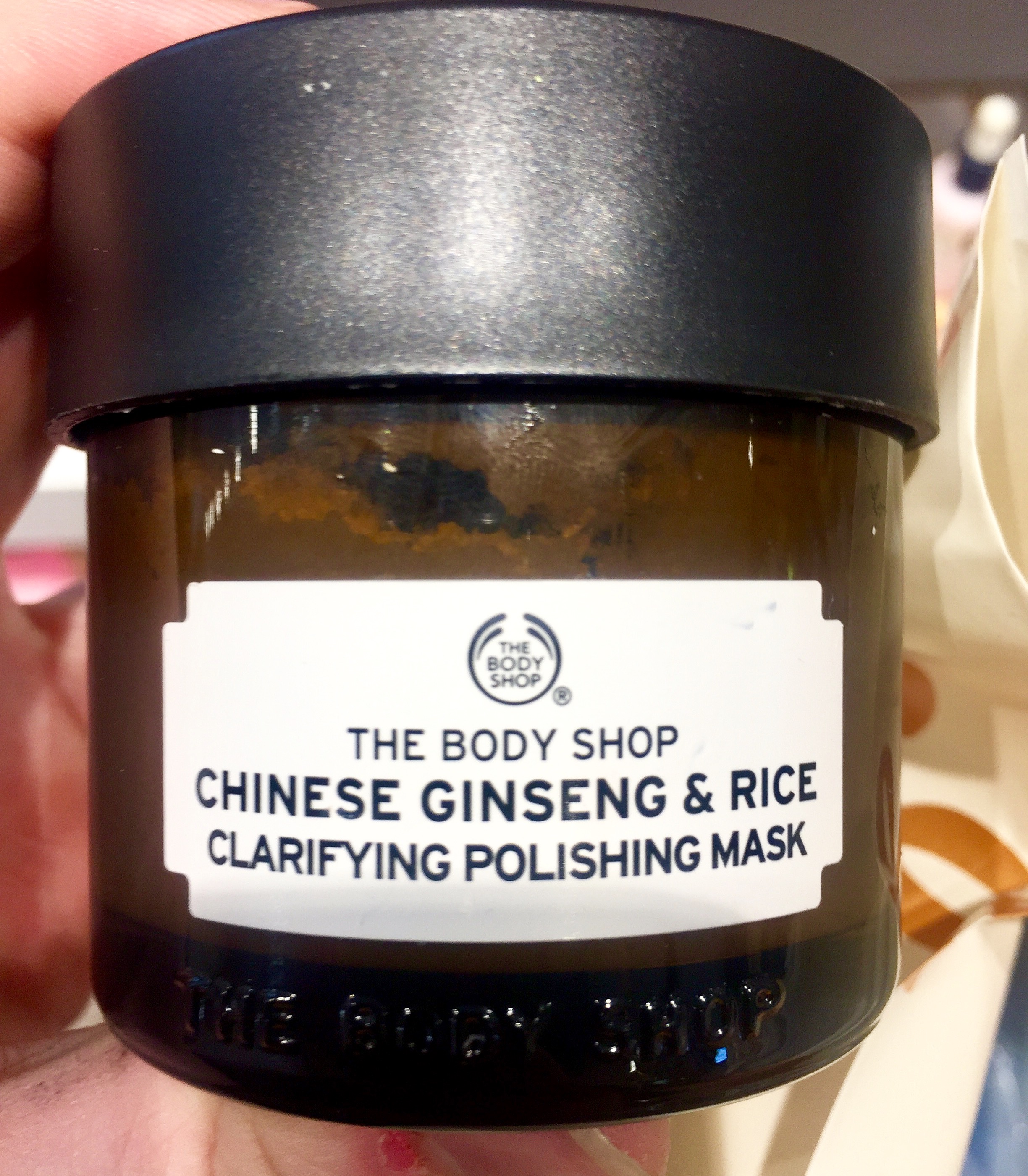 A wide black tub with a black lid and a white rectangular label that has the body shop Chinese ginseng and rice clarifying polishing mask written on it, on a light background.