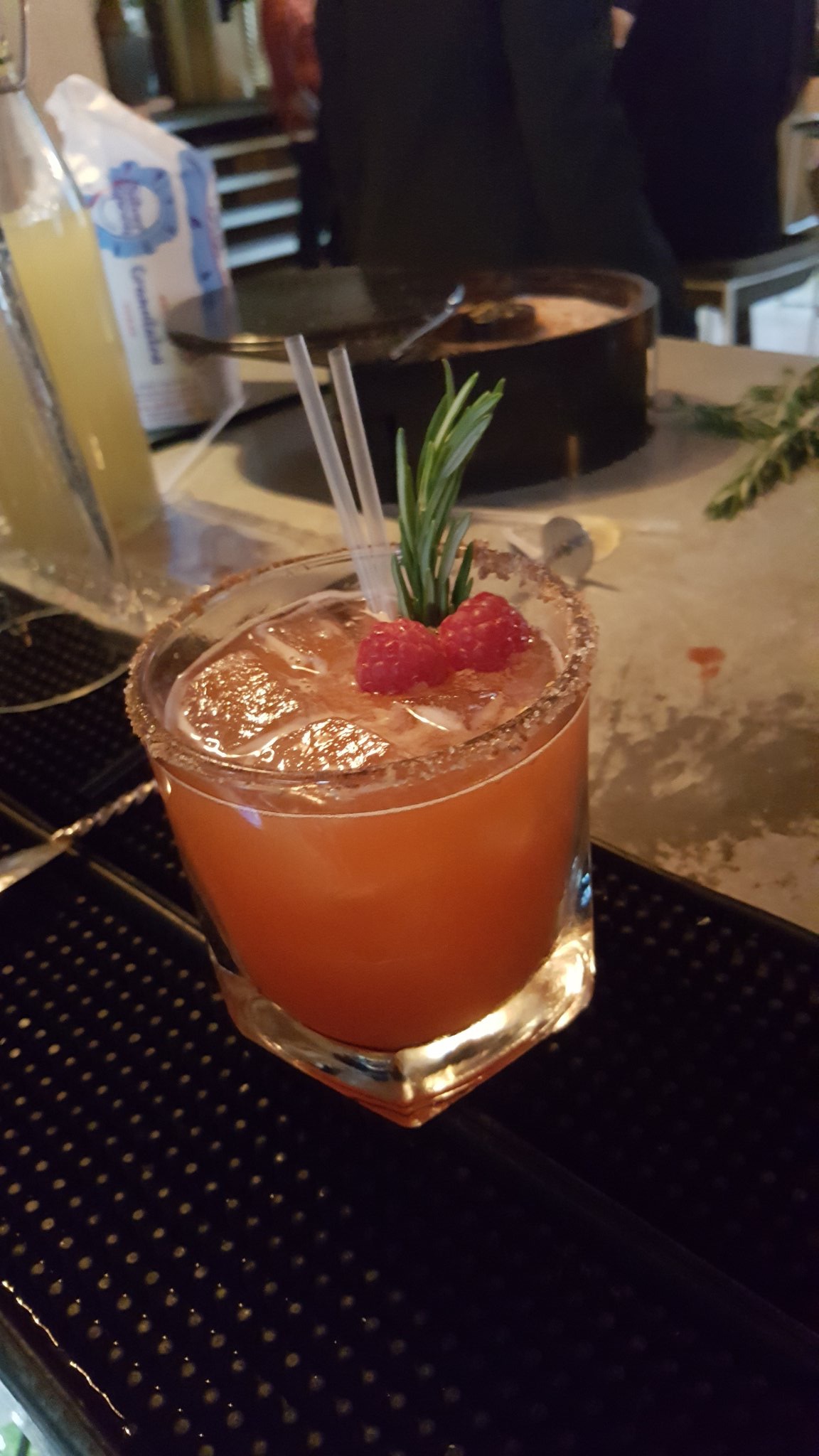 A short cylindrical glass with a sugar rim full of some pink coloured liquid and a sprig of rosemary, on a bright background.