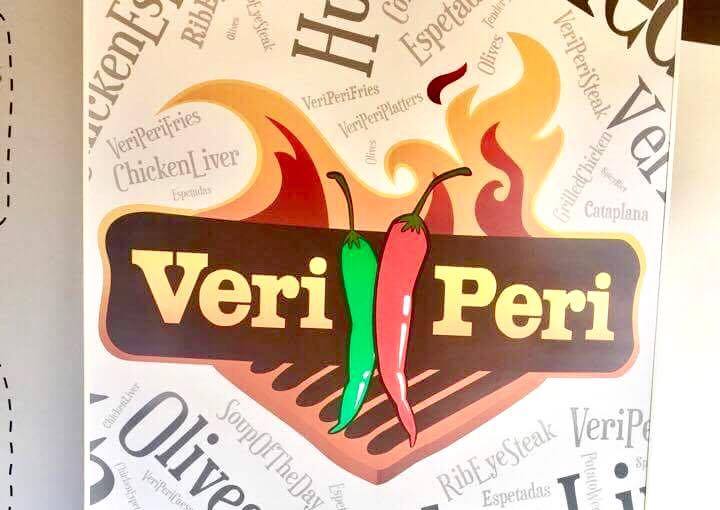 A bright white wall with a logo of fire, chillies, a purple rectangular strip and Veri Peri written in bold yellow writing and some black cursive writing below it, on a white background.