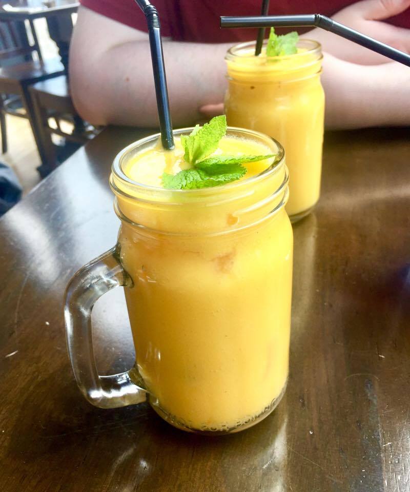 A bright yellow drink in a tall chunky mason jar with some green pieces of mint and a black straw on a dark wood table, on a dark background.