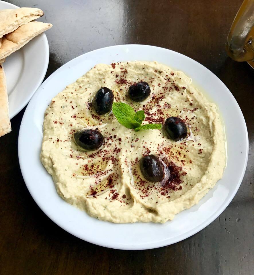 Some pale thick bright beige hummus with some green herbs and some black pitted olives inside a white small low bowl, on a dark background.