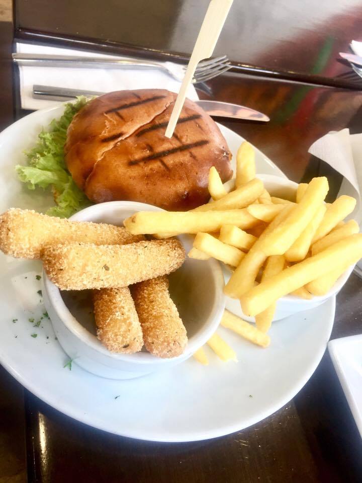 A medium circular bun with some grill marks on it filled with a dark brown Beef Patty and some yellow cheese and a wooden stick going through it next to some circular pots filled with Fries and Mozzarella Sticks on a large circular plate on a wood table, on a white background.