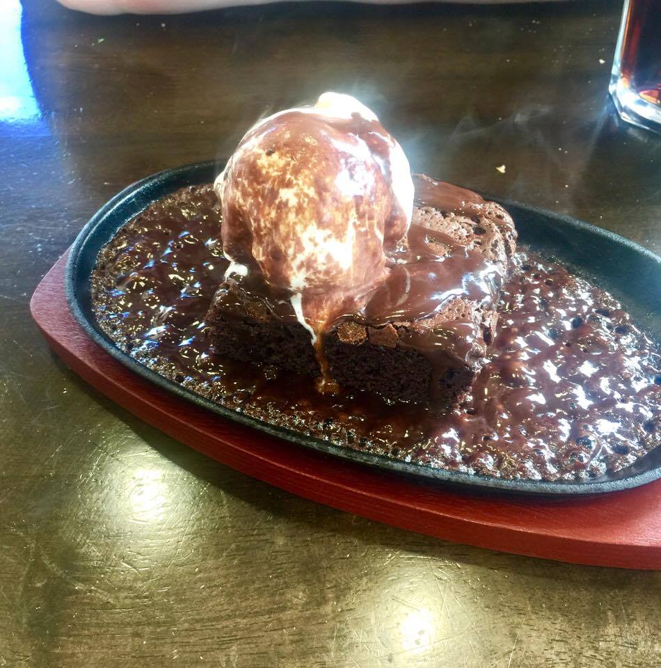A square brown chocolate brownie drenched in some bubbling light brown chocolate sauce with a circular ball of vanilla ice cream inside a brown skillet, on a dark background.