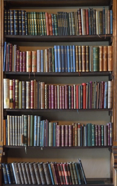 A large wide dark brown wooden bookcase filled with some thin books with different coloured covers, on a dark background.