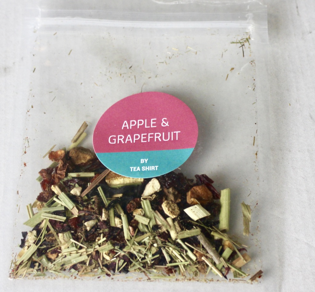 A square clear plastic bag containing some dark brown tea leaves with a light pink circular sticker that has Apple and Grapefruit written in medium white writing and Tea Shirt Tailored Refreshments written in smaller white writing on it, on a white background.