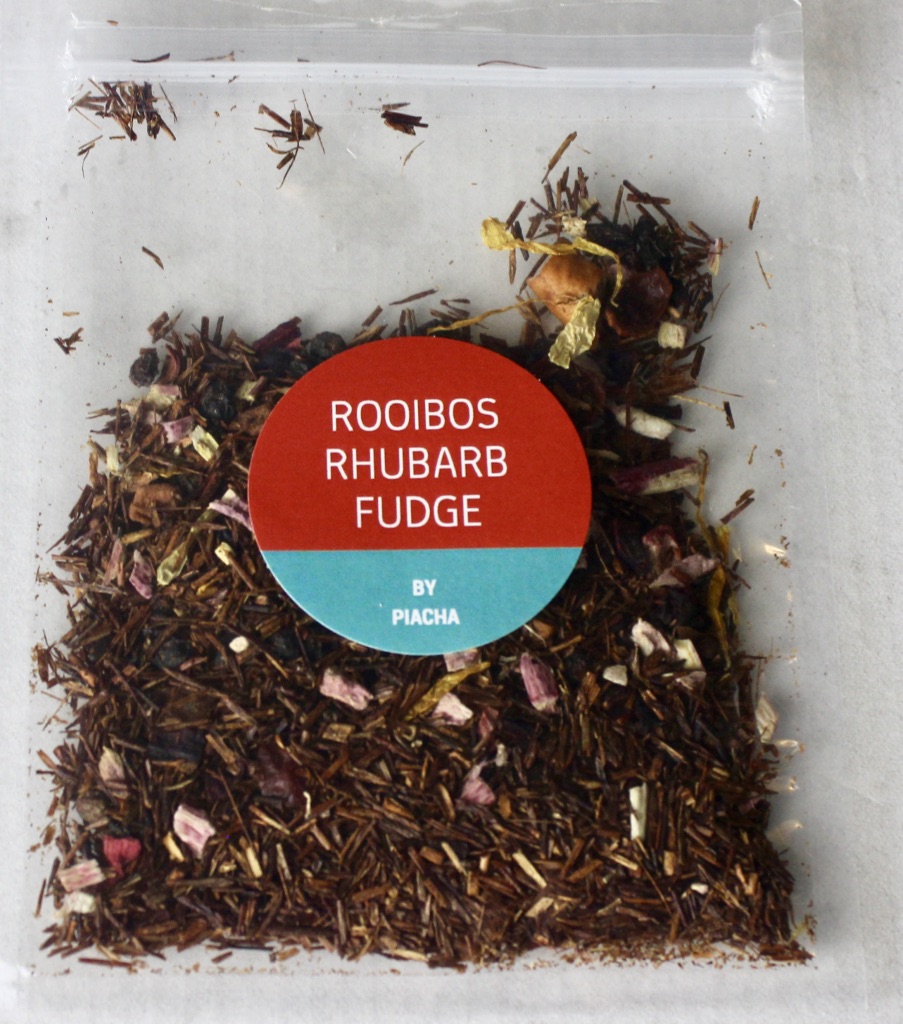 A square clear plastic bag containing some dark brown tea leaves with a dark pink circular sticker that has Rooibos Rhubarb Fudge written in medium white writing and Piacha written in smaller white writing on it, on a white background.