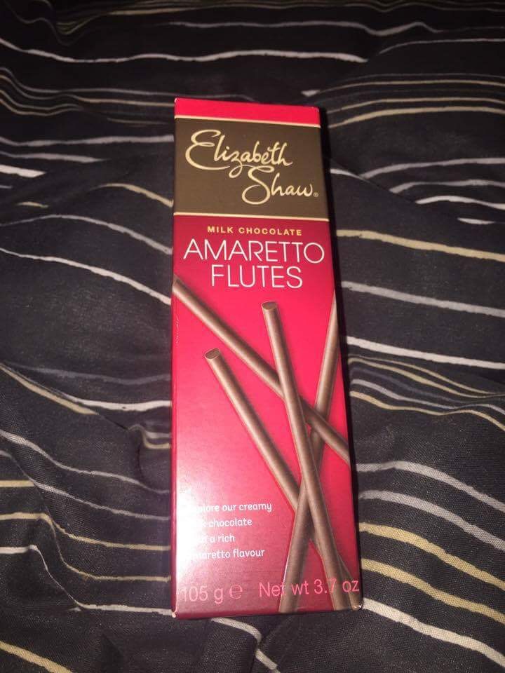 A tall rectangular box with a black strip running along the top of it that has Elizabeth Shaw written in cursive writing and amaretto flutes written in smaller writing with a picture of the chocolates next to it, on a dark background.