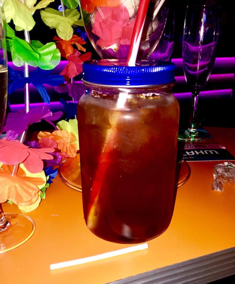 A clear mason jar with a blue screw top lid full of some red coloured liquid and a bright yellow straw, with some brightly coloured flowers in the background, on a dark background.