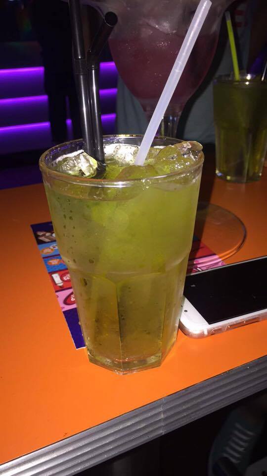 A large tumbler glass full of some light green coloured liquid, some clear ice cubes, some black straws and a glow stick, next to a white iPhone on a light wooden table, on a dark background.