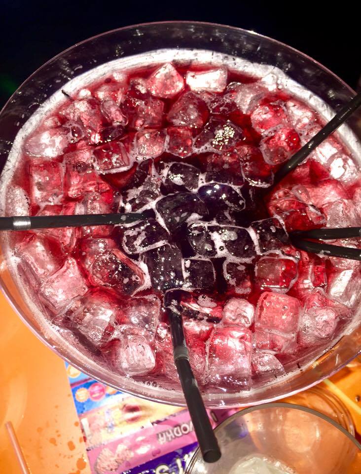 A large Martini glass full of some deep purple liquid, some clear ice cubes and some black straws, on a light wooden table, on a dark background.