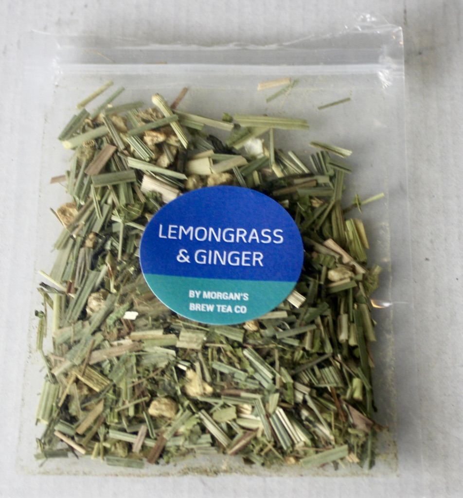 A square clear plastic bag containing some dark brown tea leaves with an electric blue circular sticker that has Lemongrass and Ginger written in medium white writing and Morgan's Brew Tea Company written in smaller white writing on it, on a white background.