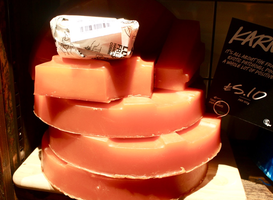 Some half circles of bright Pink coloured soap stacked on top of each other with one covered in bright white paper that has Lush written all over it, on a dark background.