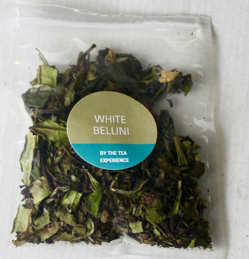 A square clear plastic bag containing some dark brown tea leaves with a green circular sticker that has white Bellini written in medium white writing and the tea experience written in smaller white writing on it, on a white background.