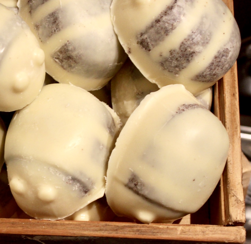 Some bright yellow blocks of moisturiser in the shape of some Bees with some grey stripes on them and some yellow dots for eyes in a square light wooden box, on a light background.