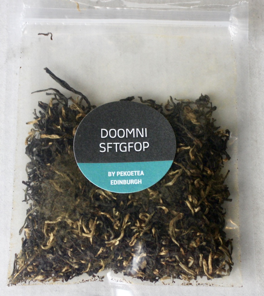 A square clear plastic bag containing some dark brown tea leaves with a black circular sticker that has Doomni Sftgfop written in medium white writing and Pekoetea Edinburgh written in smaller white writing on it, on a white background.
