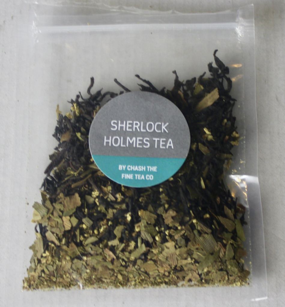 A square clear plastic bag containing some dark brown tea leaves with a light grey circular sticker that has Sherlock Holmes Tea written in medium white writing and Chash The Fine Tea Company written in smaller white writing on it, on a white background.