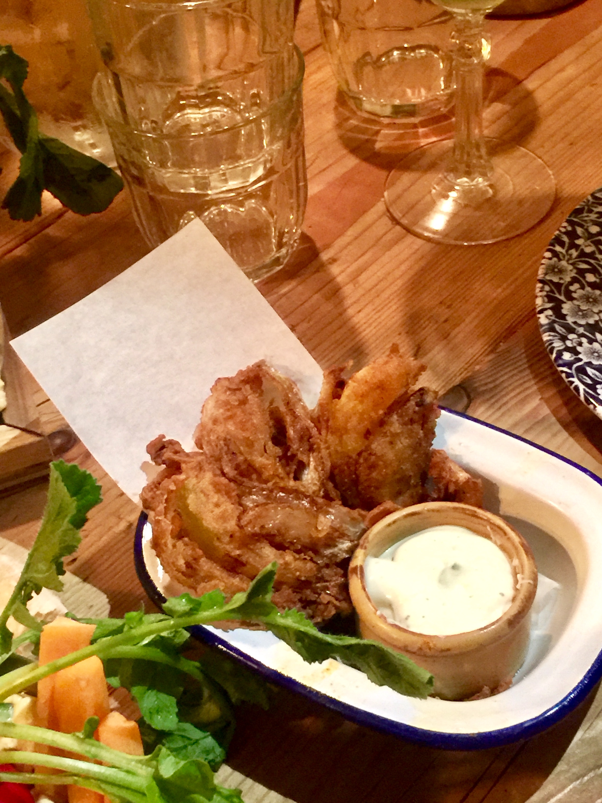 A bunch of Golden brown crispy oval onion petals next to some square white napkins and a little white circular pot of Creme Fraiche on a wooden table, on a dark background.