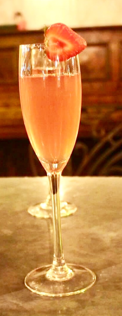 A large champagne flute full of some pale pink coloured liquid and a strawberry on the Rim of the glass on a light brown wooden table, on a dark background.