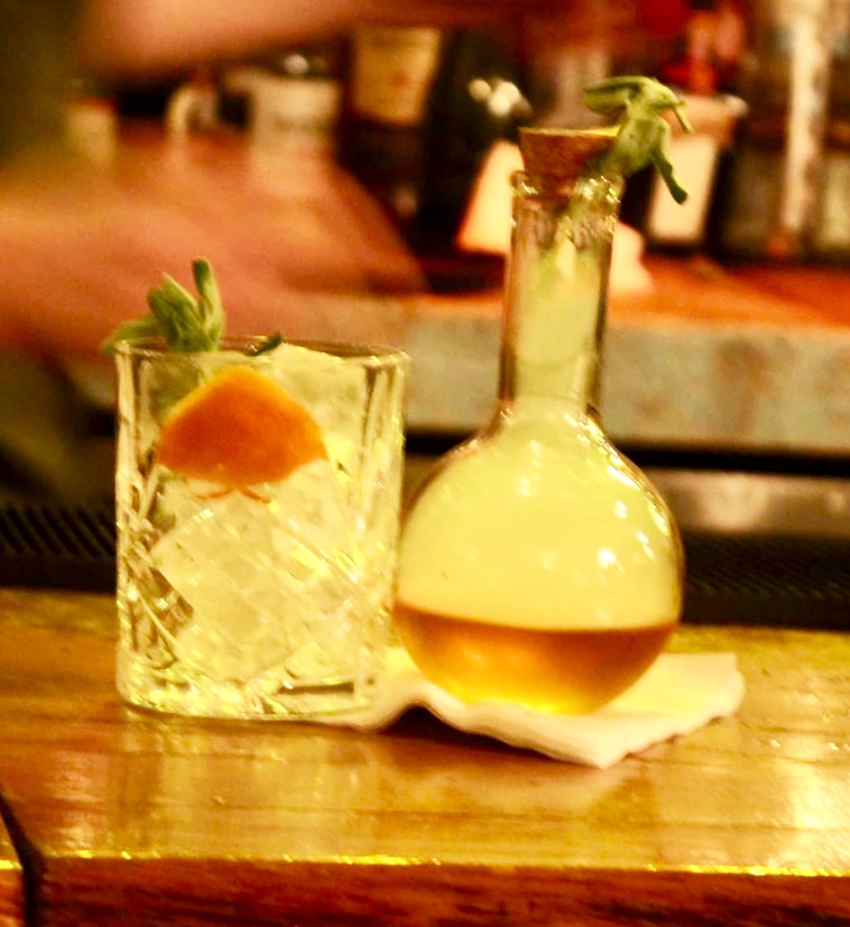 A short clear cylindrical tumbler glass full of some clear ice cubes and some orange coloured liquid next to a scientific bottle full of some bright green coloured liquid on a light wooden table, on a dark background.