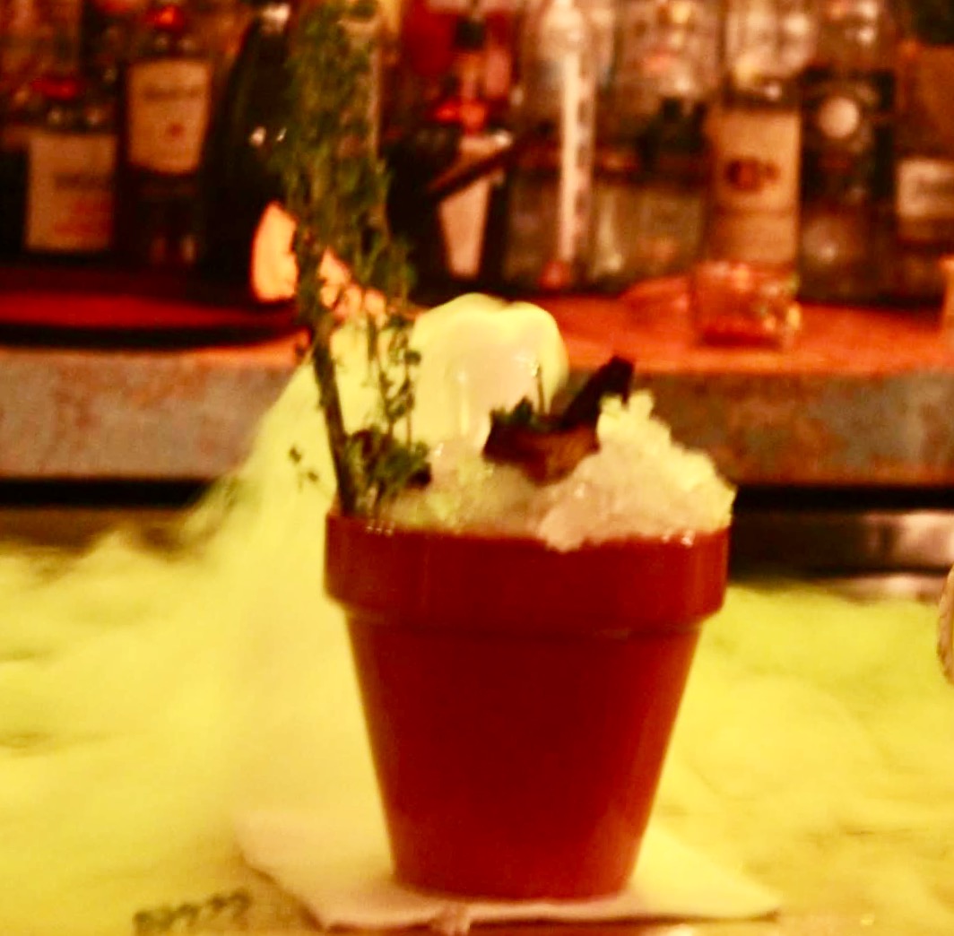 A large light wooden plant pot filled with some clear ice cubes, some black straws, and a cocktail hidden beneath some dry ice on a light wooden bar, on a dark background.