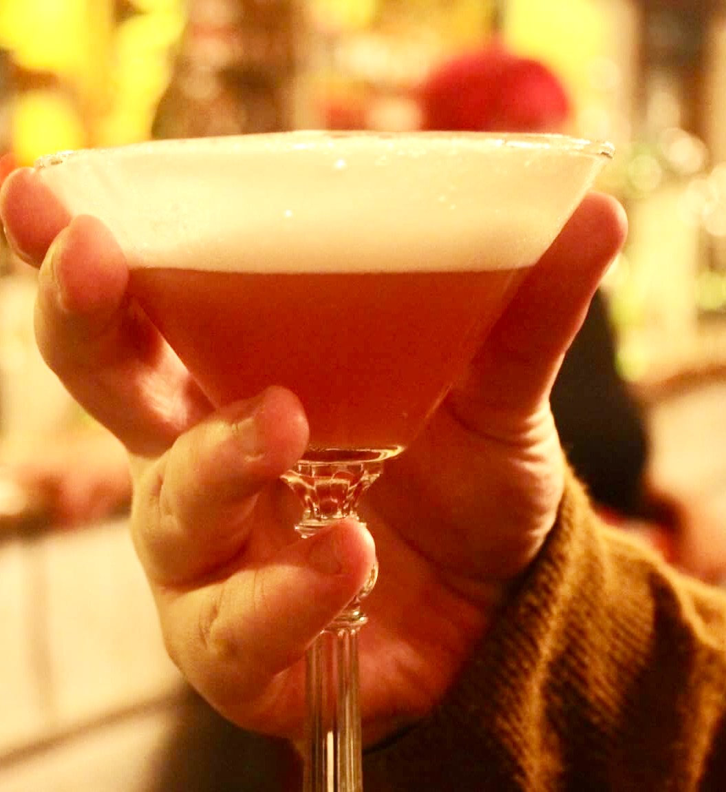 A pale pink hand holding a large stemmed Martini glass full of some deep red coloured liquid and a layer of frothy white egg white foam, on a dark background