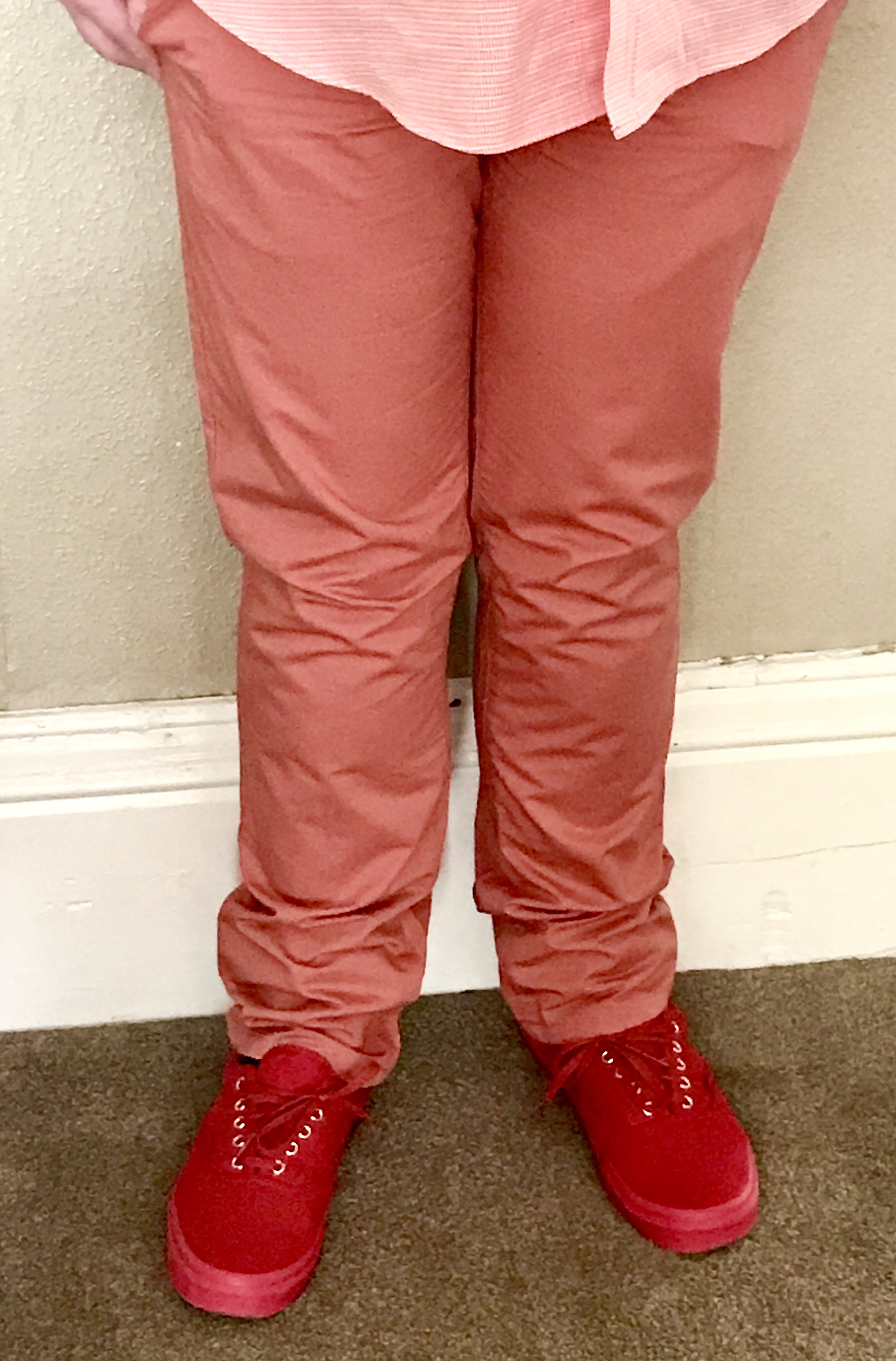 Me stood against a white wall wearing a pair of burnt orange chinos and a pair of red shoes with some red laces and soles on a green carpet, on a light background.