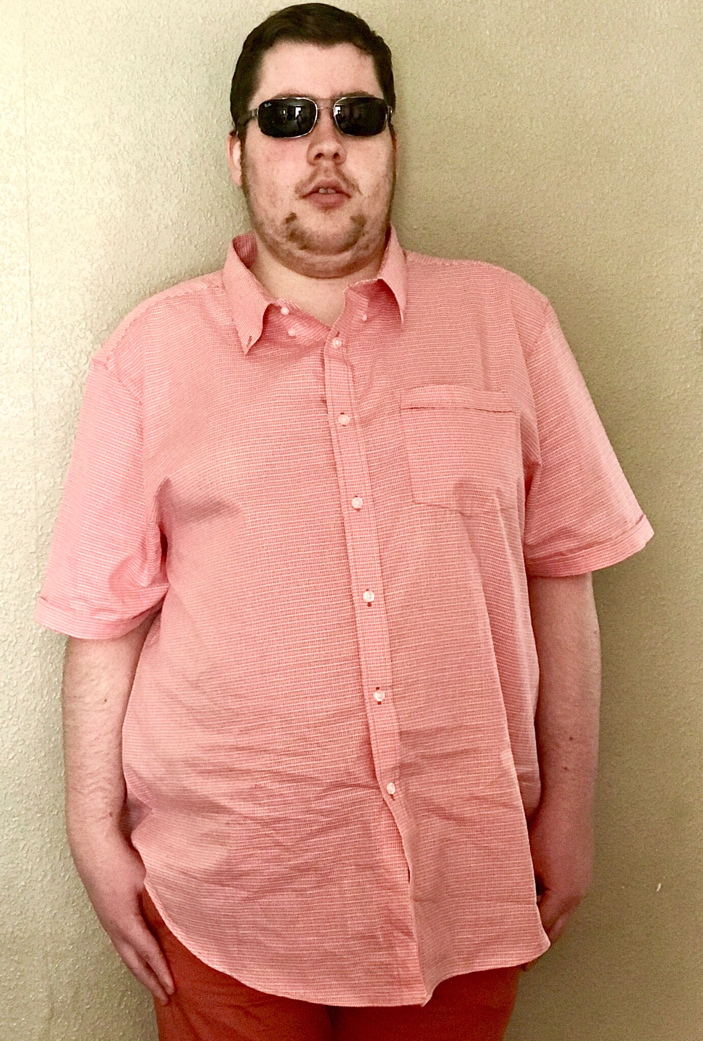 Me wearing a pair of dark green sunglasses and a bright orange shirt next to a white wall, on a bright background.