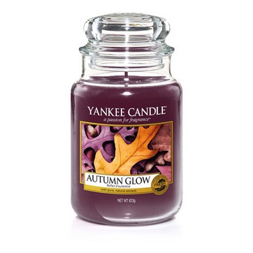 A tall glass jar full of some deep brown coloured wax with a label that has yankee candle written in white writing, Autumn Glow written in black writing, and a picture of some brown leaves on it, on a white background.