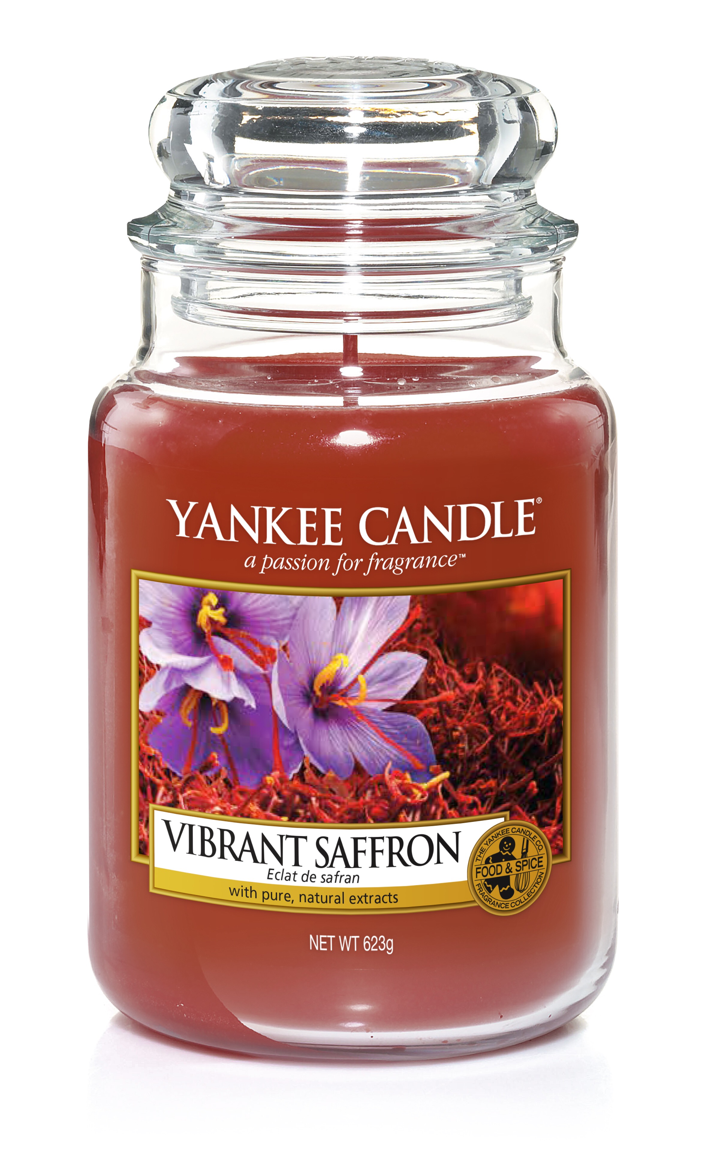 A tall glass jar full of some dark gold coloured wax with a label that has yankee candle written in white writing, Vibrant Saffron written in black writing, and a picture of some white flowers and some golden saffron on it, on a white background.