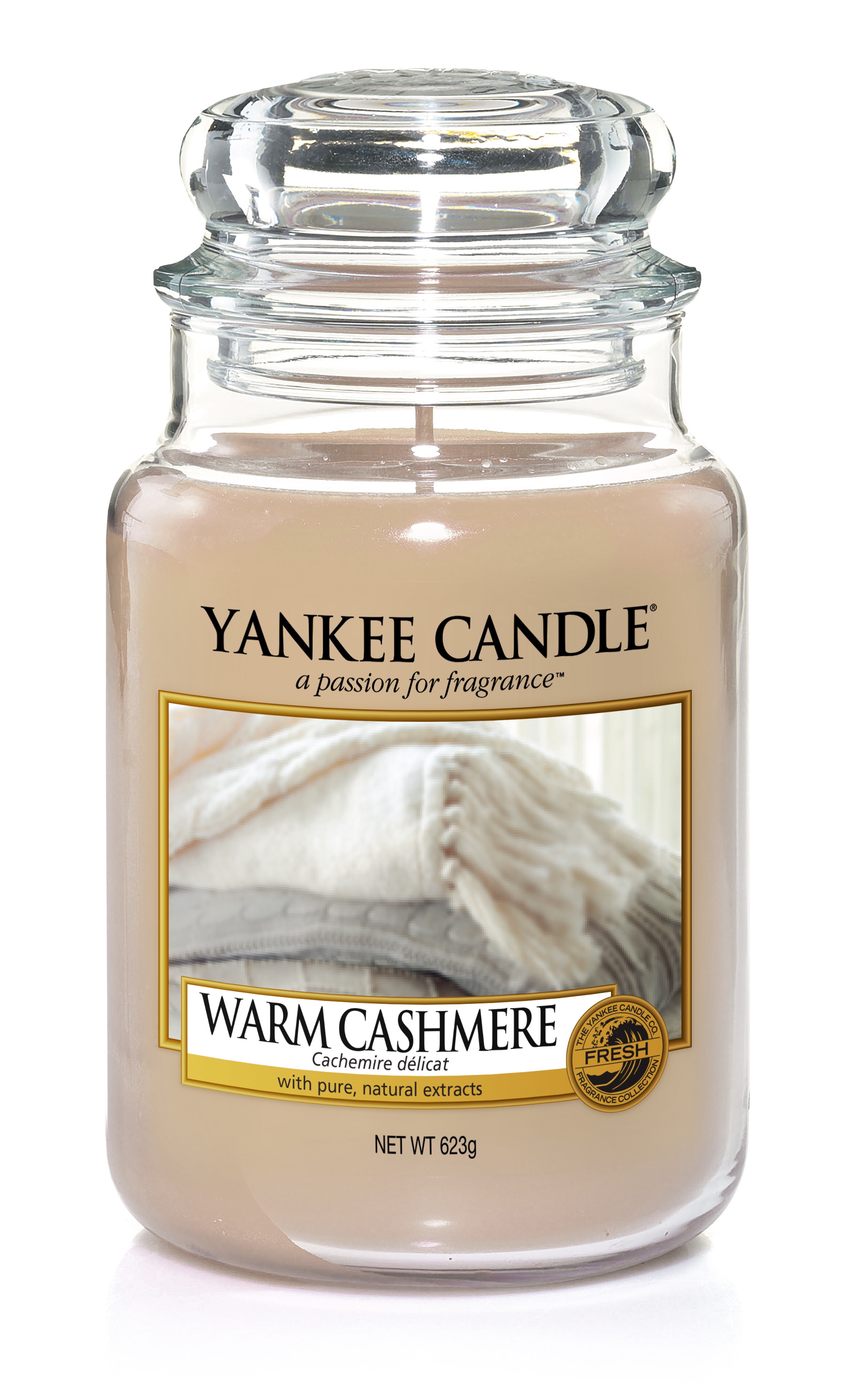 A tall glass jar full of some cream coloured wax with a label that has yankee candle written in white writing, Warm Cashmere written in black writing, and a picture of some white fabric on it, on a white background.